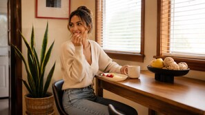 A person seated at a wooden table with a bowl of fruit and a mug, in a room with a potted plant and a window with blinds.