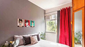 A bedroom with a bed, a nightstand, a window with red curtains, and wall art. - Boutique Apartment | 5 min from Copacabana Beach and Metro | Restaurants, Bars and Shops (Rio de Janeiro)