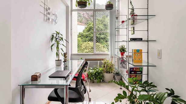 A modern office space with a glass table, office chair, potted plants, a bookshelf, and a window with a view of greenery. - Boutique Apartment | 5 min from Copacabana Beach and Metro | Restaurants, Bars and Shops (Rio de Janeiro)