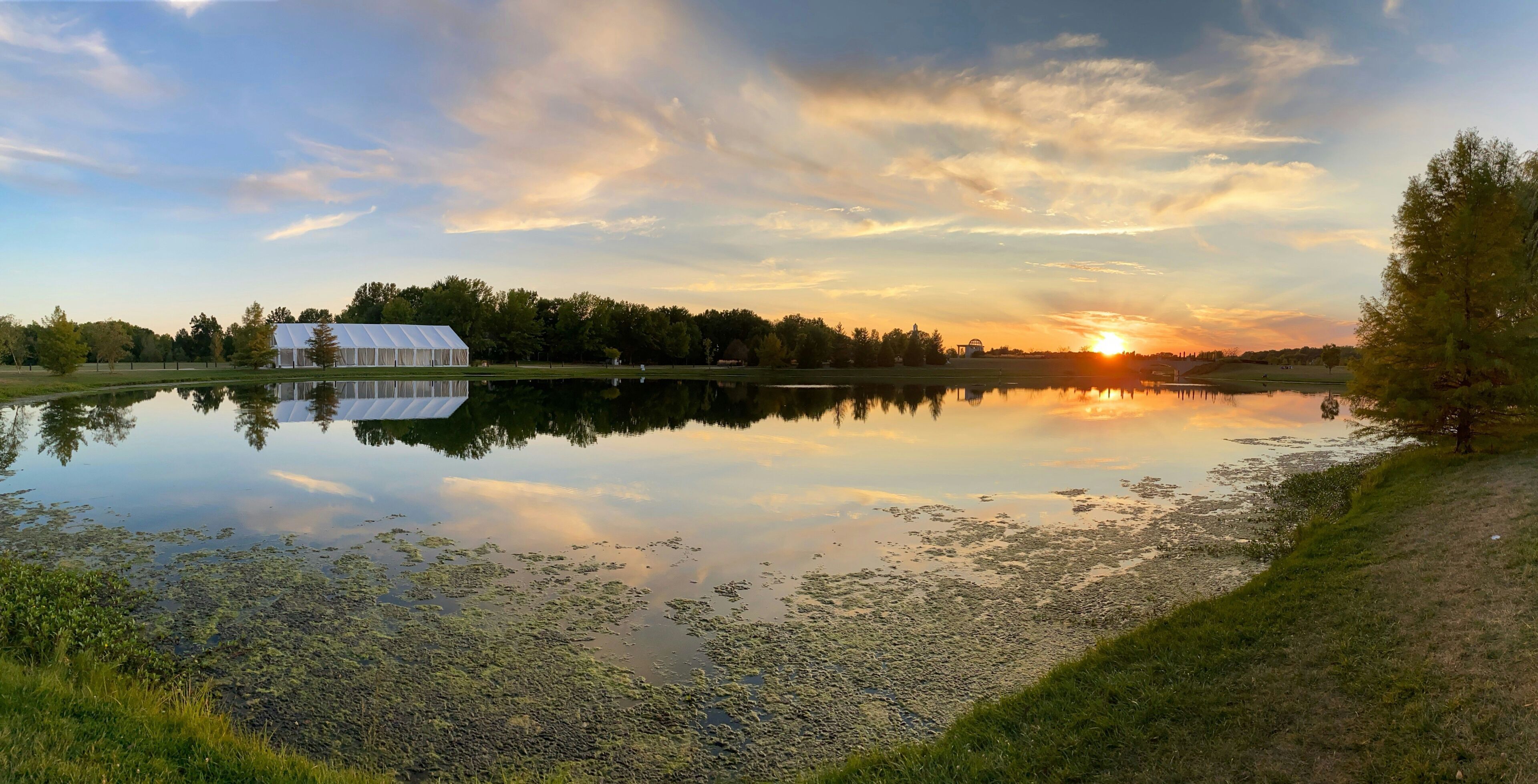 Una vista serena su un lago, con una struttura a forma di tenda bianca, acqua calma e alberi che si riflettono al tramonto.