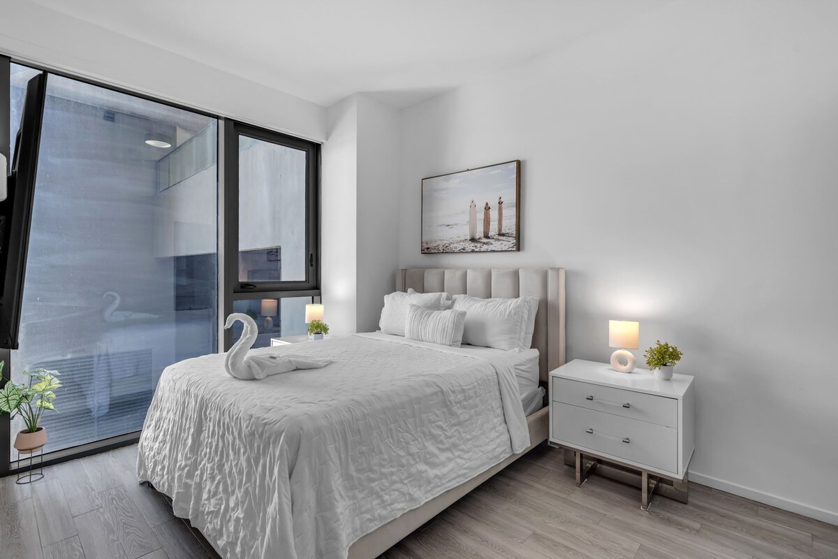 A modern bedroom with a large bed, bedside tables, a wall-mounted picture, and a view through a glass door.