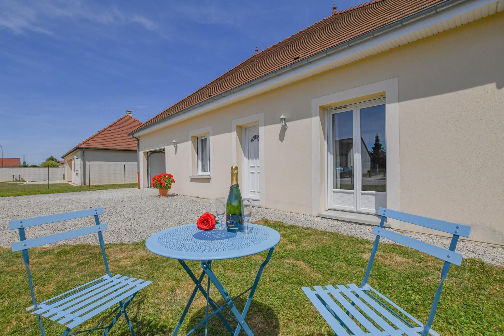 A house with a gravel driveway, a front door, and a window. There is a table with a bottle and glasses, and two blue chairs.