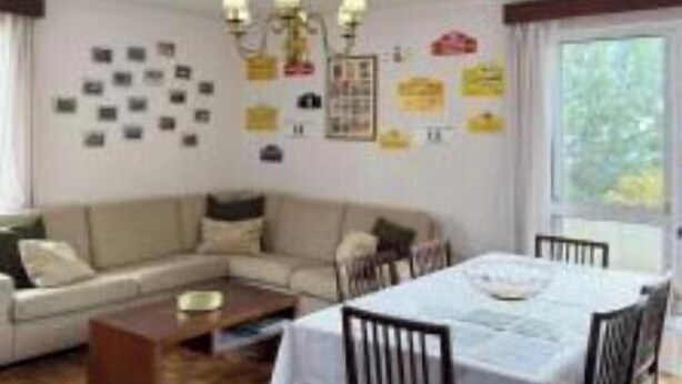 A living room with a sofa, coffee table, dining table, and chairs. There is a chandelier, a window with curtains, and a wall with various pictures and notes. - Funchal Petrolhead Apartment (Funchal)