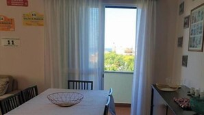 A room with a dining table, chairs, a window with curtains, a dresser, and a view of greenery outside. - Funchal Petrolhead Apartment (Funchal)