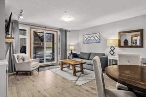 A living room with a sofa, armchair, wooden coffee table, dining table, and chairs. - Cozy Retreat- Gym, BBQ, Free Parking (Canmore)