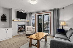 A living room with a fireplace, a sofa, a coffee table, a TV mounted on the wall, and a sliding glass door leading to a balcony. - Cozy Retreat- Gym, BBQ, Free Parking (Canmore)
