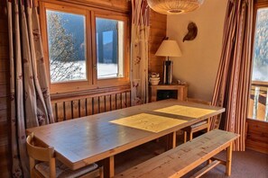 Ein Holzblockhaus-Interieur mit einem großen Esstisch, Bänken, einer Lampe und Blick auf die verschneite Landschaft durch die Fenster.