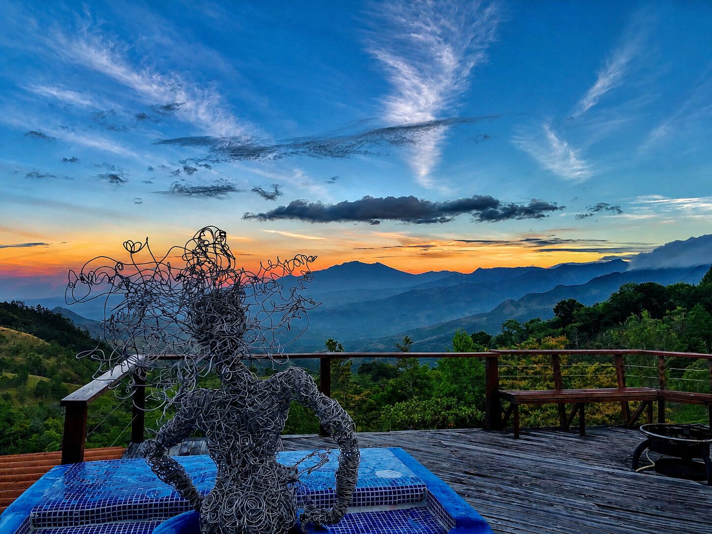 A sculpture of a human figure on a wooden deck with a view of mountains and a sunset.
