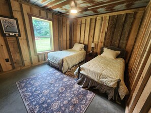 A rustic room with two single beds, a small table with a lamp, a window with a view of greenery, and a framed picture on the wall. - Yellowhammer Lodge near Lake Weiss and Little River Canyon (Gaylesville)