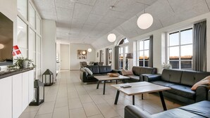 Un vaste espace de vie comprenant un canapé, une table basse et une table à manger. De grandes fenêtres, équipées de rideaux, donnent sur l’extérieur et on peut y apercevoir un drapeau.