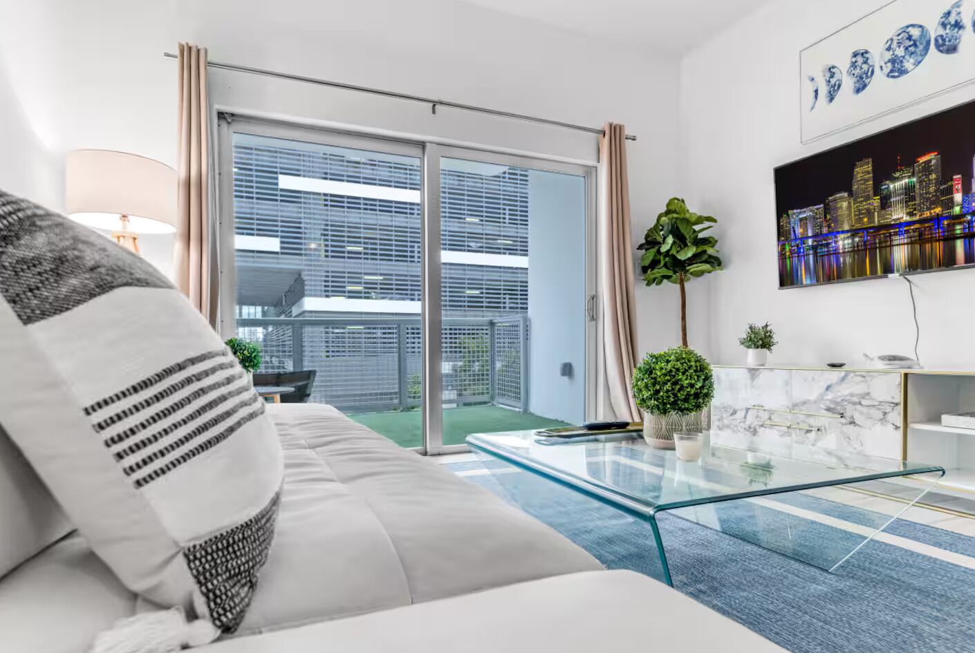 A modern living room with a glass coffee table, a sofa with patterned cushions, a potted plant, and a cityscape artwork on the wall.