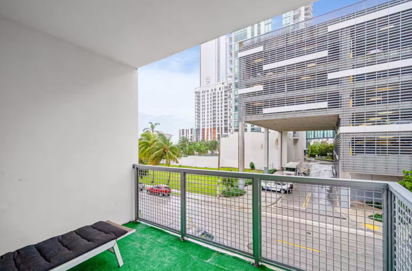 A balcony with a green artificial turf floor, a metal railing, and a bench. In the background, there are modern buildings, a palm tree, and a street with cars.