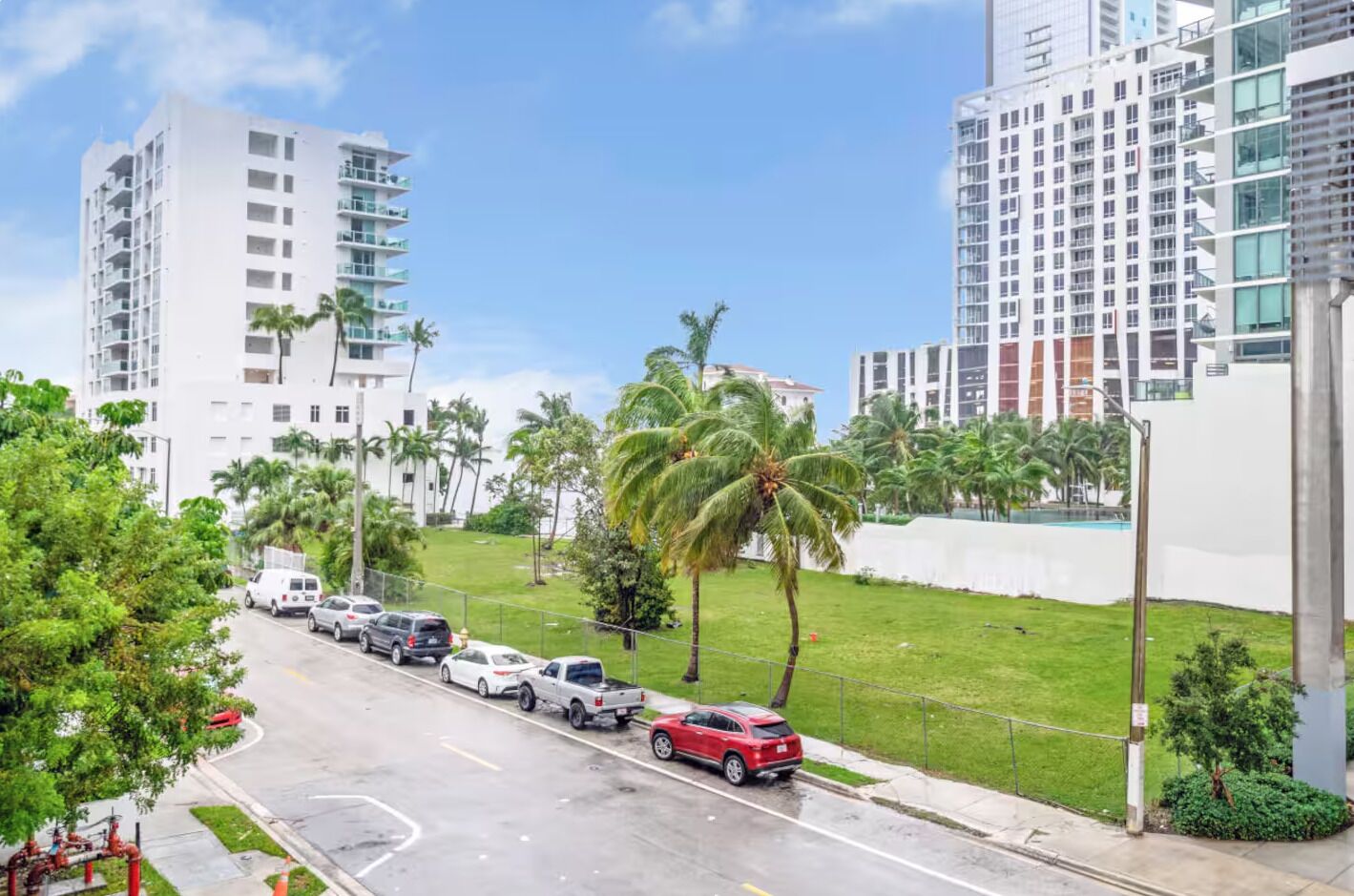 A street view with modern buildings, a grassy area with palm trees, and parked cars.