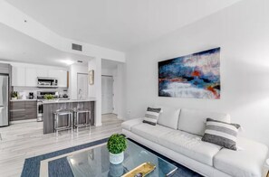 A modern kitchen with stainless steel appliances, a dining area with a glass coffee table, and a living room with a white sofa.