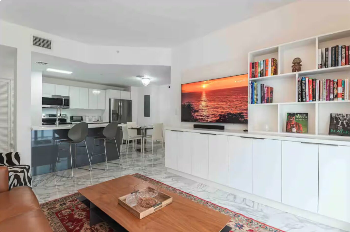 A modern living room with a flat-screen TV, a bookshelf, and a dining area with chairs.
