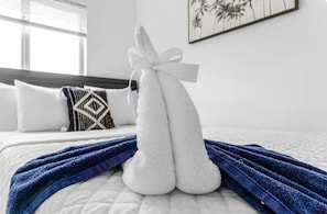 Una cama bien tendida con una toalla blanca y una toalla azul dobladas encima, un cojín estampado, y una pared con un cuadro de una palmera.