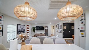 Un salon moderne avec un coin repas, comprenant une grande table blanche, deux suspensions en osier et un téléviseur à écran plat fixé au mur.