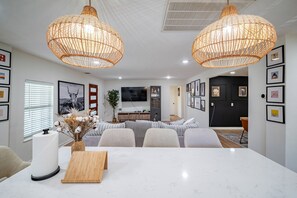 A modern living room with a dining area, featuring a large white table, two wicker pendant lights, and a flat-screen TV mounted on the wall. - Modern Comfort: Spacious 6-Bedroom, 5-Bath Home (Houston)