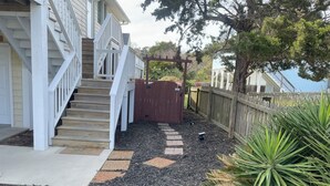 Eine Holzterrasse mit einer Treppe zu einem Haus, ein eingezäunter Bereich mit einem Tor und ein Pfad mit trittfesten Steinen.