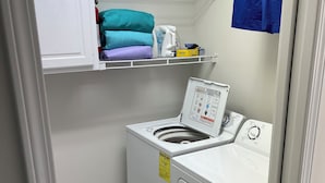 Ein Waschraum mit einer Waschmaschine mit Frontlader, einem Regal mit gefalteter Kleidung und einem blauen Handtuch an der Wand.