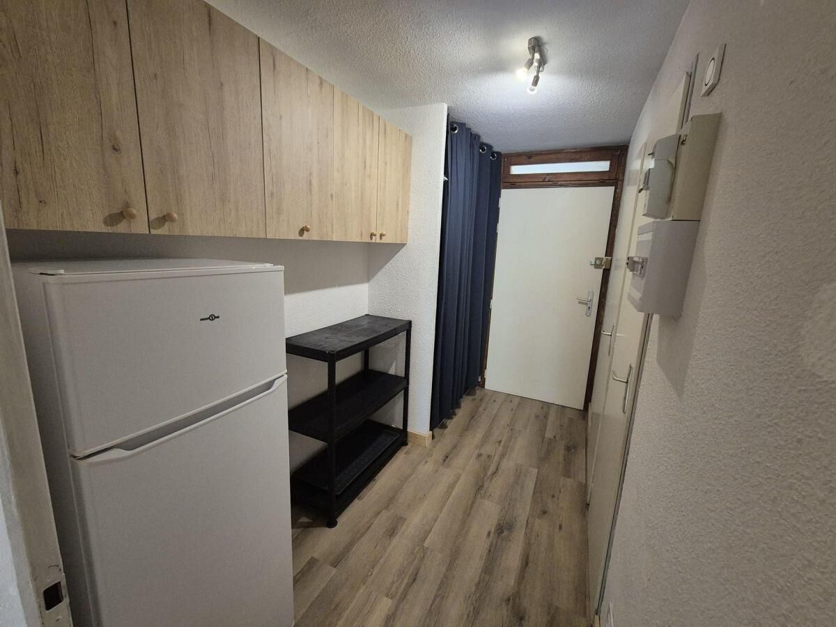 Una cocina compacta con armarios de madera, un refrigerador blanco, una estantería negra y una cortina oscura.
