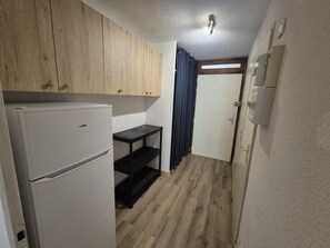 A compact kitchen with wooden cabinets, a white refrigerator, a black shelf unit, and a dark curtain. - 100m from ski slopes, Ground floor, terrace, tv, ski locker, 20m², Valloire (Valloire)