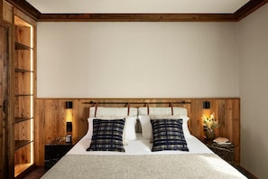 Ein Schlafzimmer mit Holz-Kopfteil, einem Bett mit weißen Leinen, zwei gemusterten Kissen, einem Nachttisch mit Lampe und einem Nachtschränkchen.