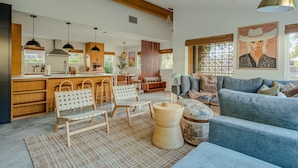 A spacious living area with a blue sofa, wooden chairs, a woven basket, and a kitchen with wooden cabinets and pendant lights. - Casa Tranquila • Pool, Ping Pong, Game Room & More (Austin)