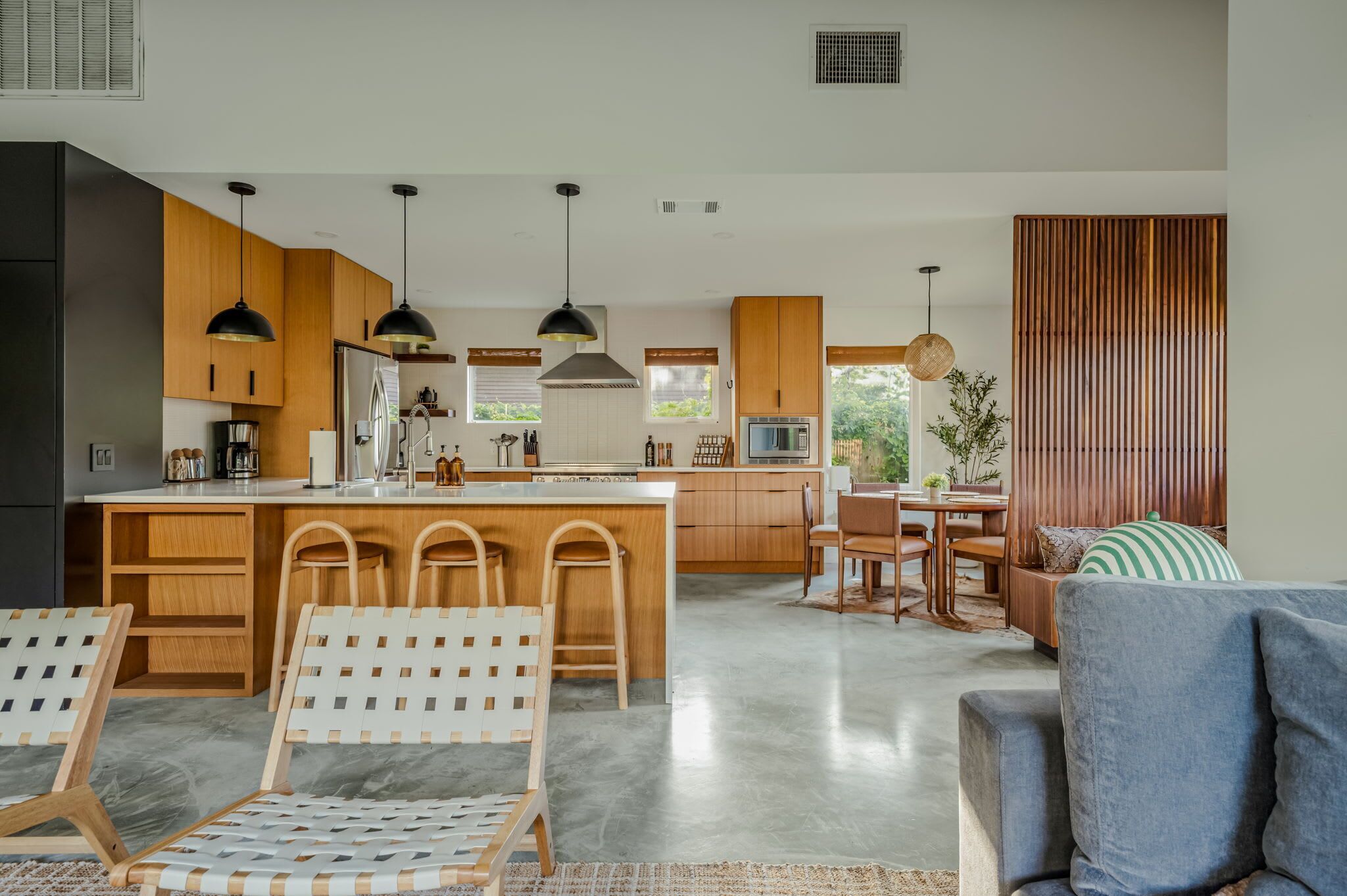 A modern kitchen with wooden cabinets, a central island, bar stools, pendant lighting, and a dining area with a wooden table and chairs.