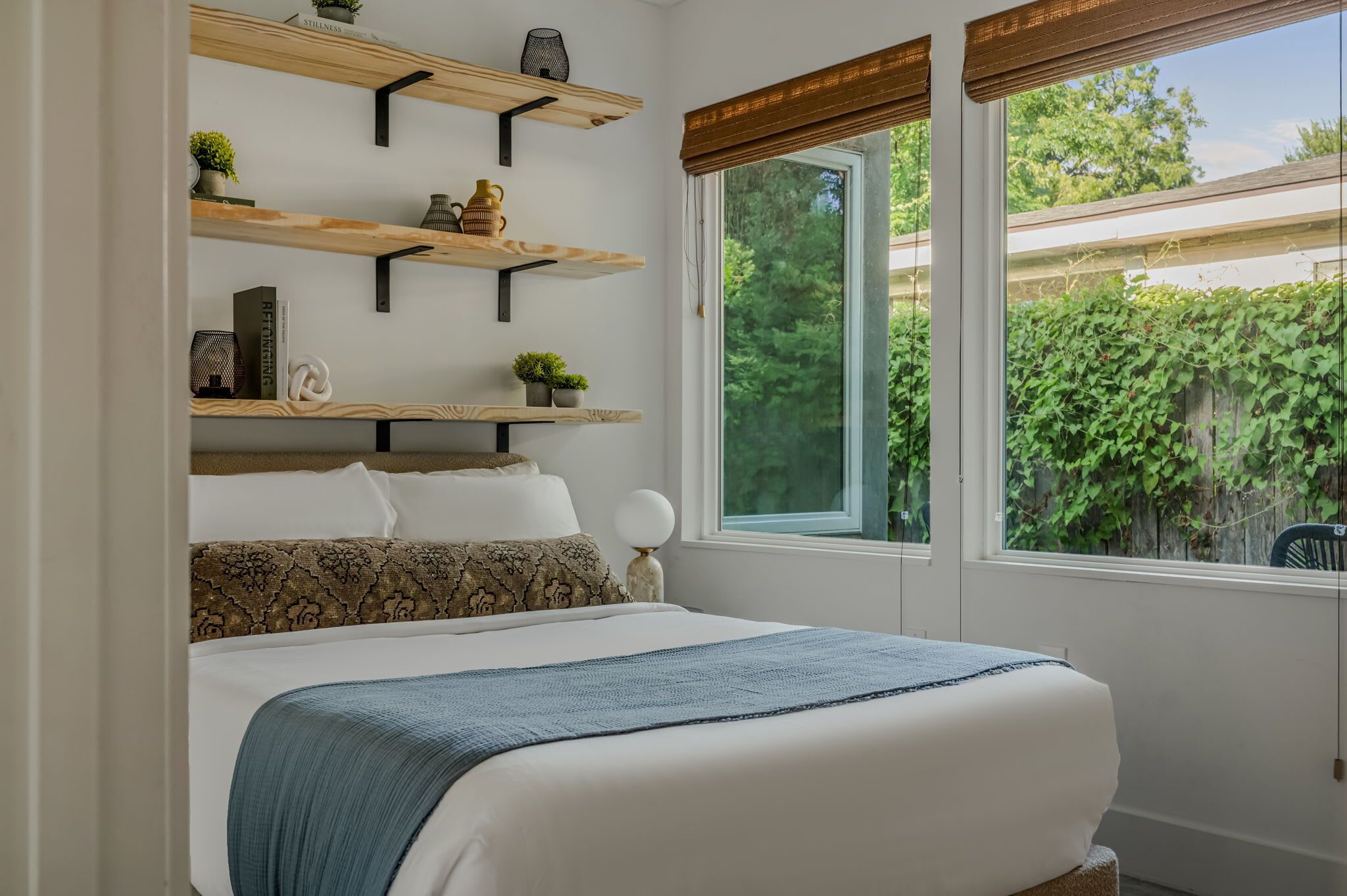 Un dormitorio con cama, una ventana con vistas a zonas verdes y estanterías de madera con objetos decorativos.