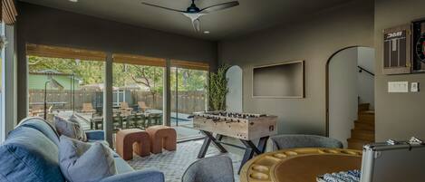A modern living room with a foosball table, comfortable seating, and a large window overlooking a backyard.