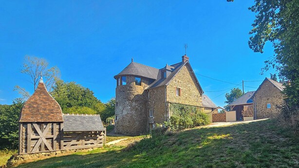 Un bâtiment en pierre avec un toit à forte pente, une grange en bois et un ciel d’un bleu limpide.