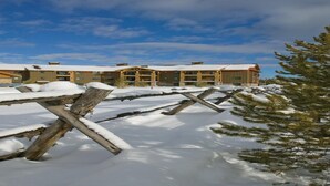 Un espacio exterior cubierto de nieve, con una cerca de madera y un edificio con múltiples ventanas y un techo rojo al fondo.