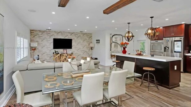 A modern kitchen with a central island, a dining area with a glass table, and a living area with a sofa and TV.