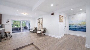 A spacious room with a wooden floor, two wicker chairs, a bench, a large abstract painting, and a view through glass doors.