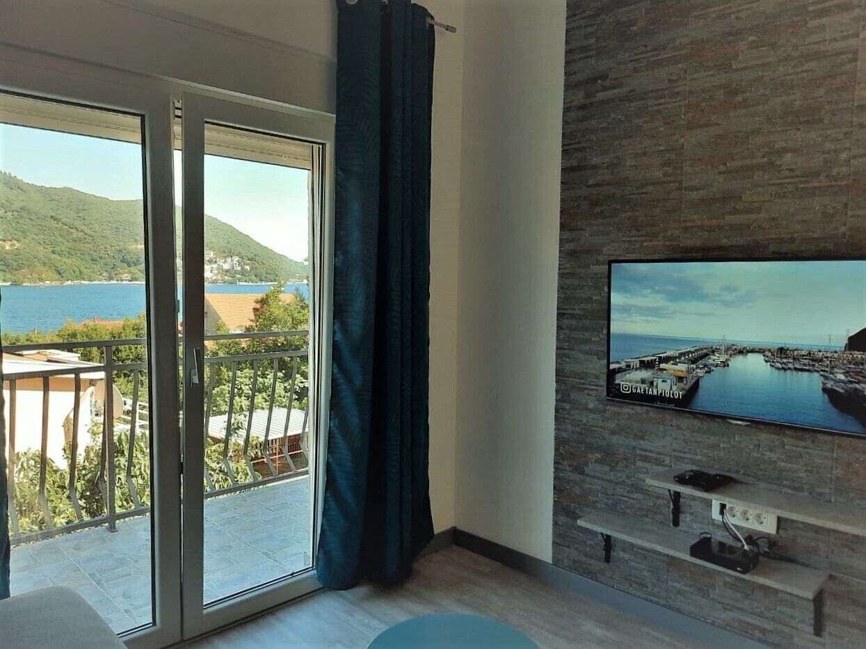 A room with a TV mounted on a brick wall, a sliding glass door to a balcony, and a view of a coastal area.