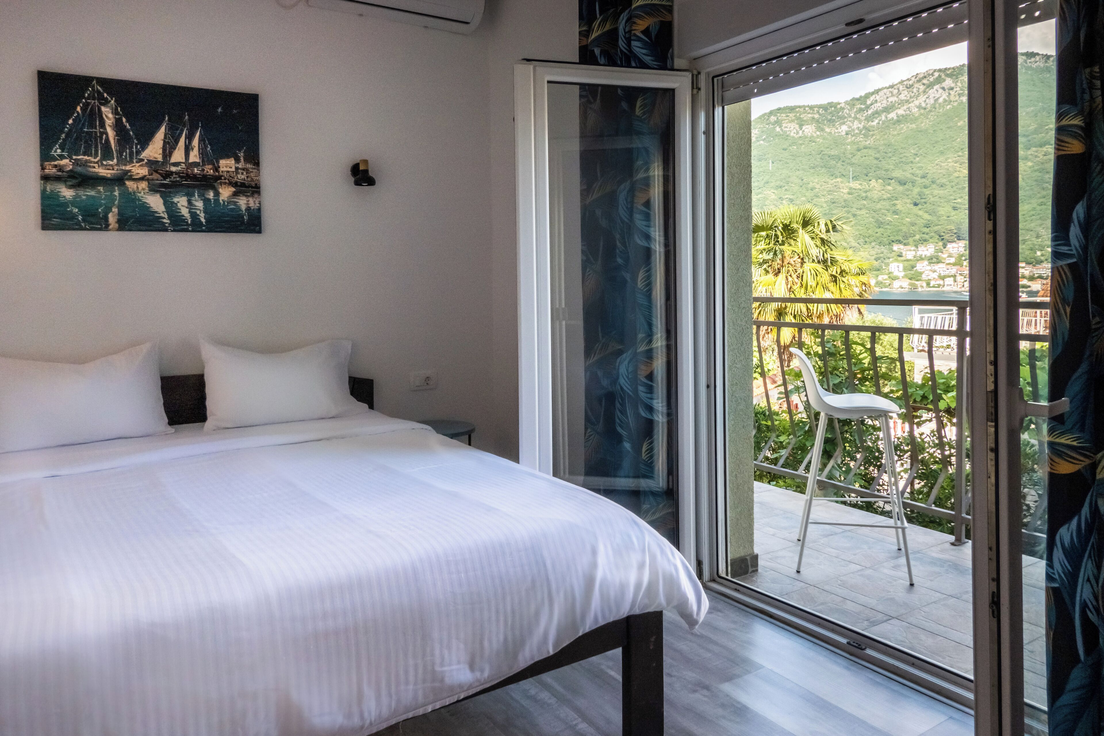 Ein Schlafzimmer mit einem Bett, einem Nachttisch, einem Gemälde und einer Schiebetür, die auf einen Balkon mit einem Stuhl führt.