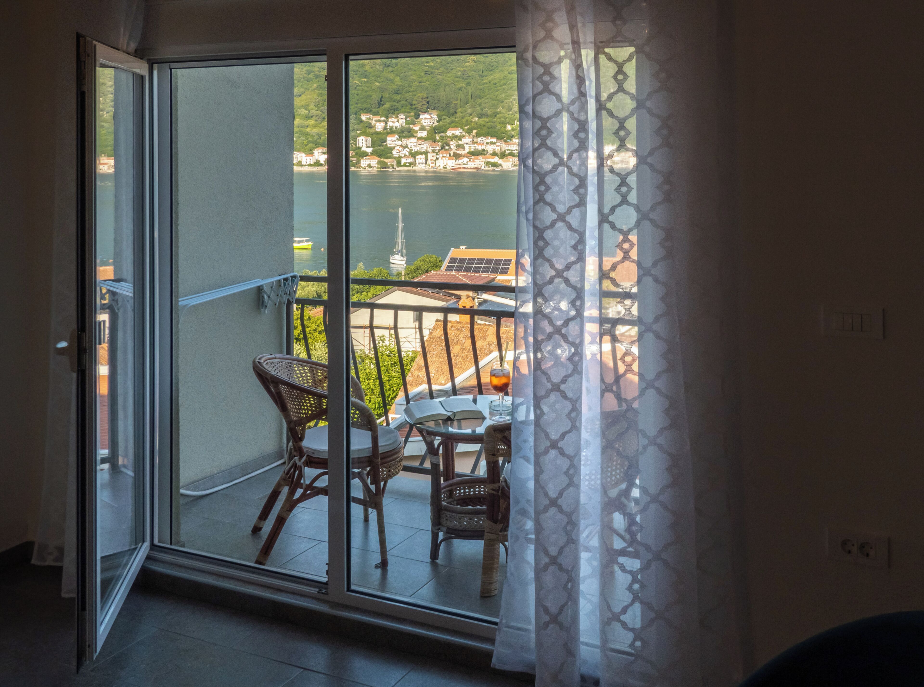 Un balcone con sedie in vimini, un tavolo e una porta vetrata che si apre su una vista panoramica di un lago e degli edifici.