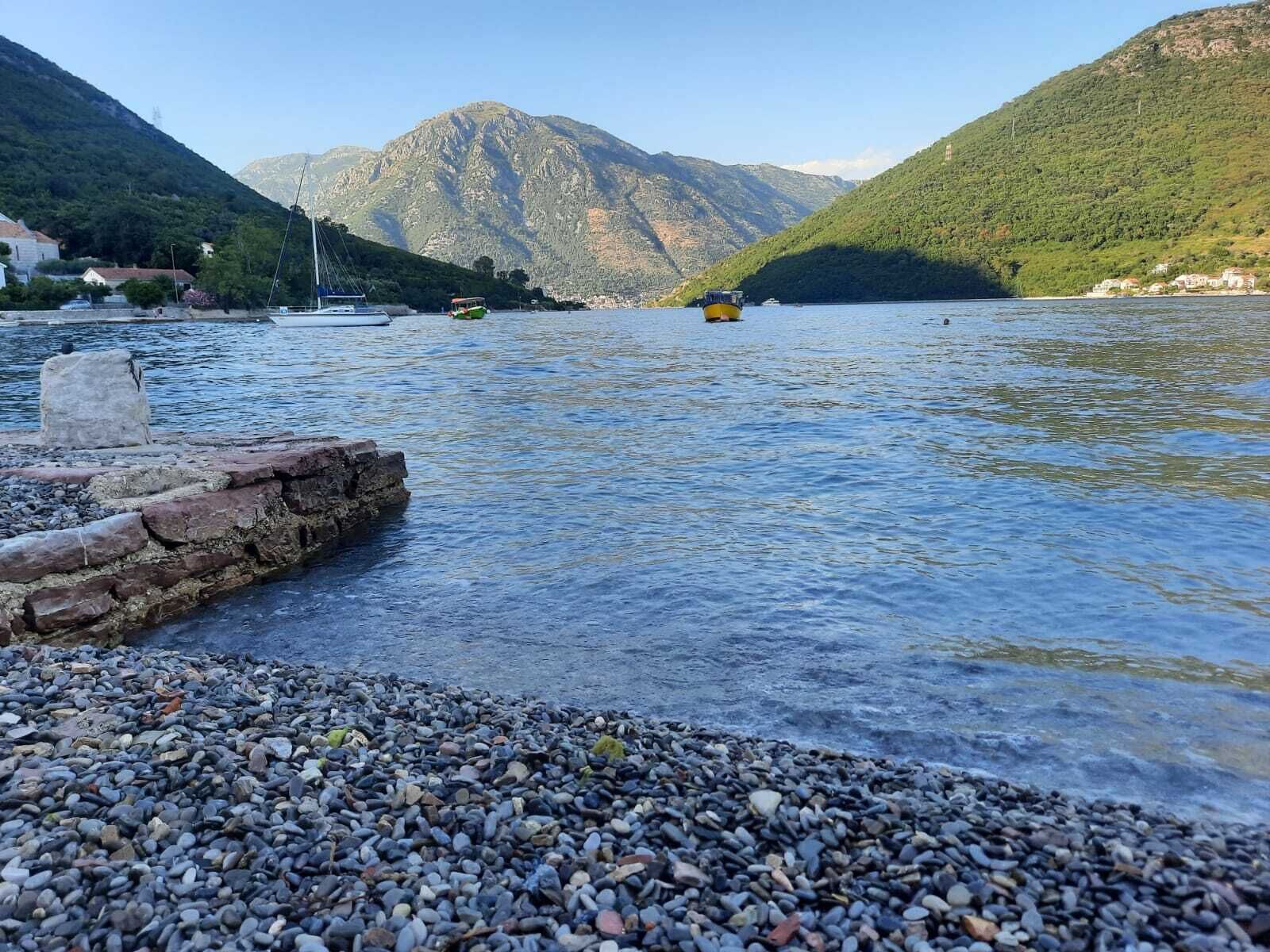 A pebbled waterfront with boats, a stone structure, and mountains in the background.