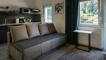 Un salon moderne comprenant un canapé, une petite table, un coin cuisine avec un four à micro-ondes et une fenêtre donnant sur un espace verdoyant.