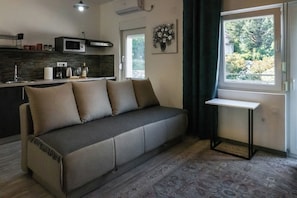 Een moderne woonkamer met een bank, een klein tafeltje, een keukenhoek met een magnetron, en een raam met uitzicht op groen.