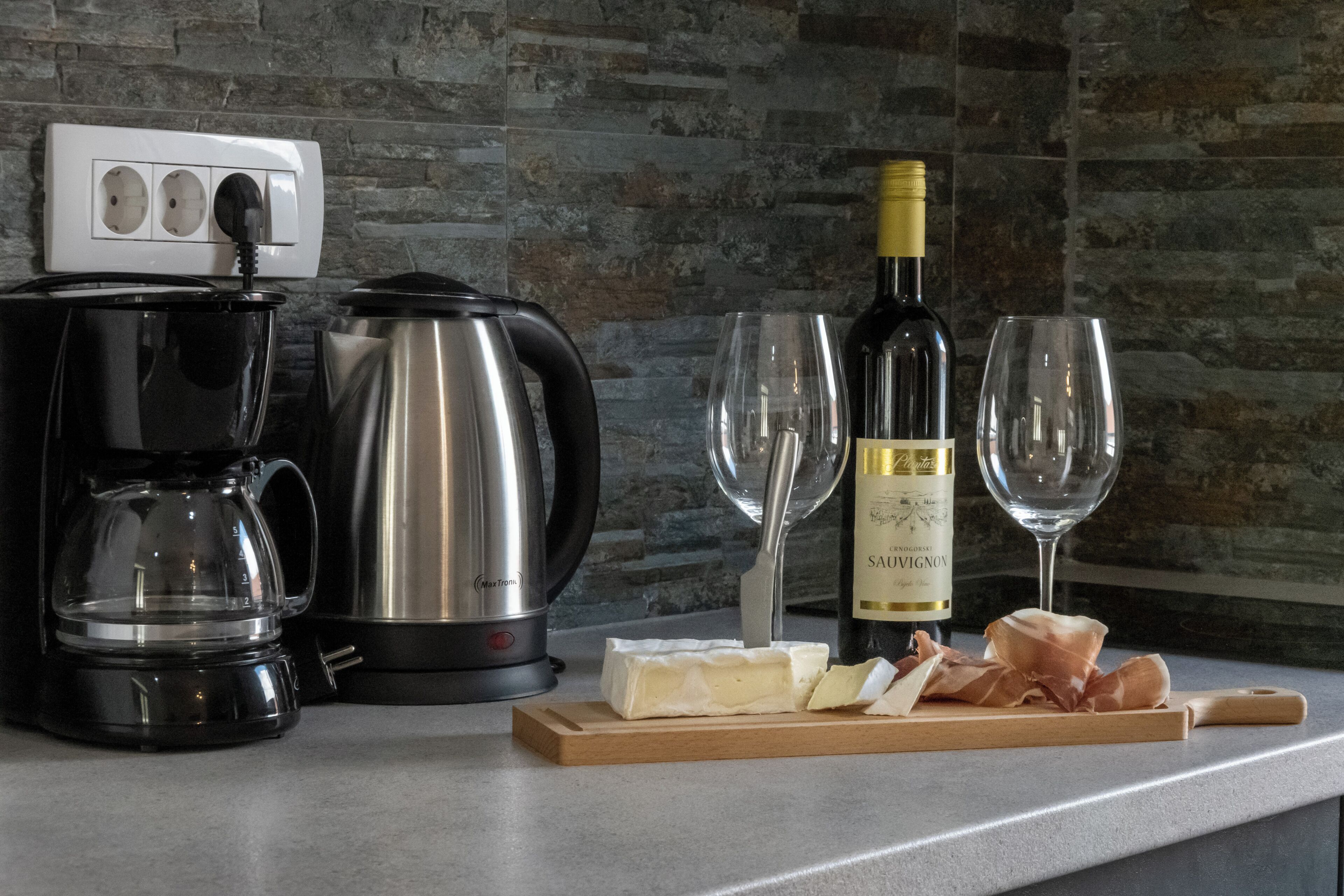 A kitchen counter with a coffee maker, a kettle, a bottle of wine, glasses, and a cutting board with cheese and meat.