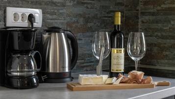 A kitchen counter with a coffee maker, a kettle, a bottle of wine, glasses, and a cutting board with cheese and meat.