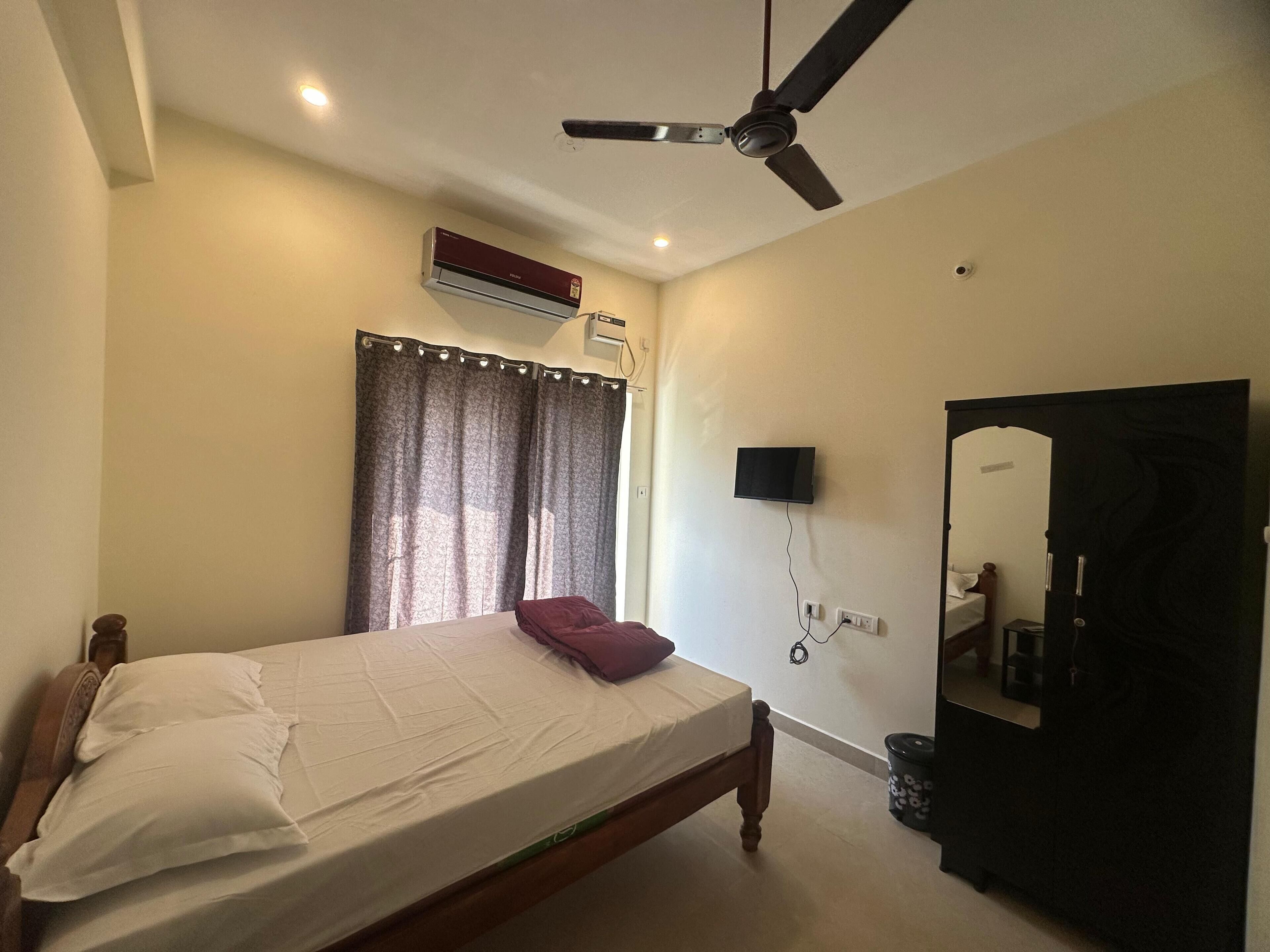 Un dormitorio con cama, ventilador de techo, unidad de aire acondicionado, televisor montado en la pared, espejo y puerta.