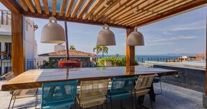 An outdoor dining area with a wooden table and chairs, a pergola with hanging lights, and a view of the sea and buildings. - Heart of Zona Romantica. Modern 1BR/2BA Oasis-Steps away to everything (Puerto Vallarta)