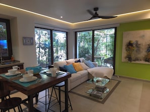 A modern living room with a sofa, dining table, and chairs, large windows with a view of greenery, and a ceiling fan. - Heart of Zona Romantica. Modern 1BR/2BA Oasis-Steps away to everything (Puerto Vallarta)