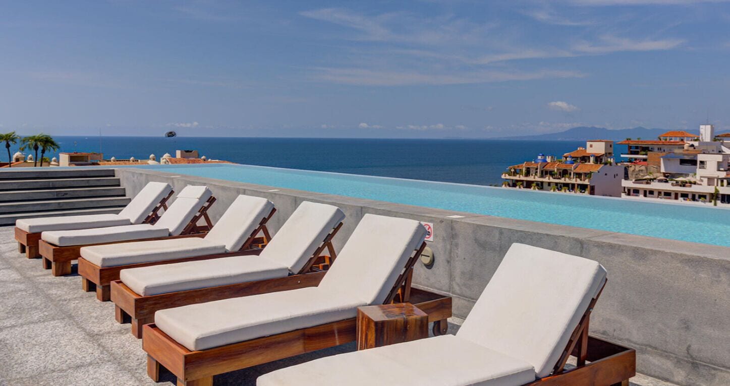 Una piscina en la azotea con sillones de descanso, vistas despejadas del océano y edificios al fondo.
