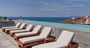 Una piscina en la azotea con sillones de descanso, vistas despejadas del océano y edificios al fondo.