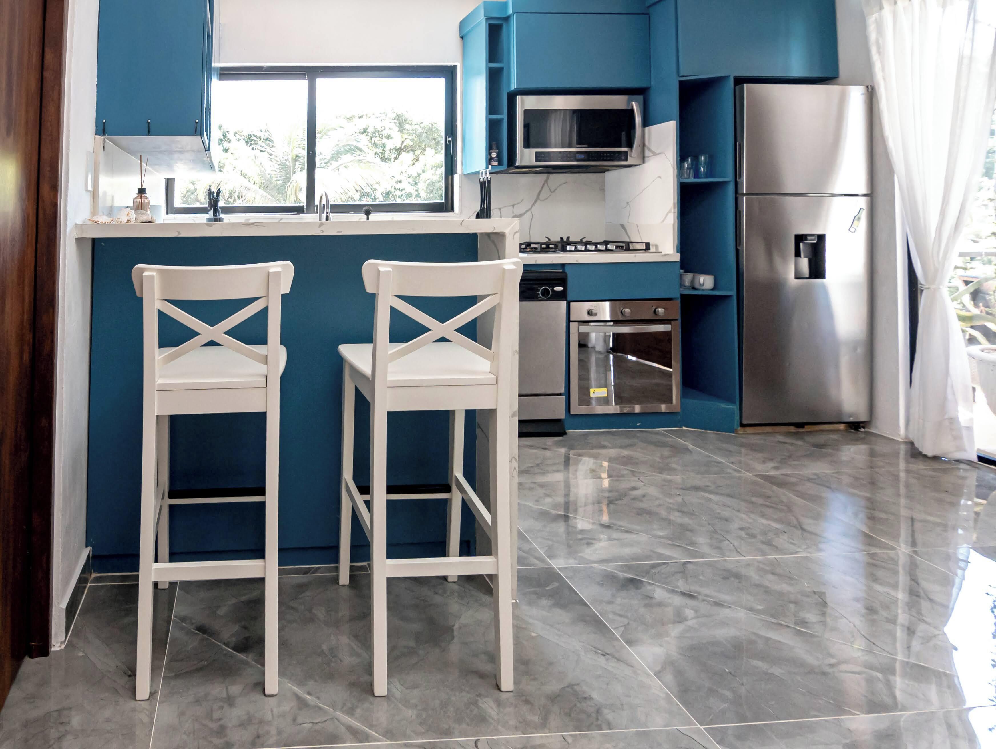 Una cocina con gabinetes azules, un microondas, un refrigerador, una estufa y dos taburetes blancos.