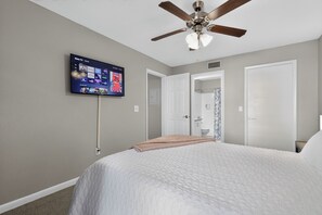 Quarto com cama, ventilador de teto, televisão na parede e porta para o banheiro.